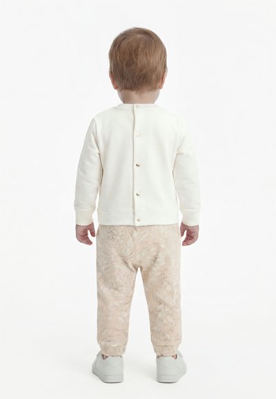 Gyermek világosbarna hajjal, háttal áll, fehér gombos pulóvert, mintás bézs nadrágot és fehér tornacipőt visel.