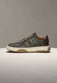 Χαμηλά sneakers με δέρμα ελιάς, καφέ μοτίβο φιδιού, μπεζ σόλα και δέσιμο μπροστά σε ανοιχτό μπεζ επιφάνεια με γκρι φόντο.