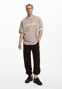 Beige oversized t-shirt med ordet "DESIGUAL" i hvid, parret med mørkebrune cargo bukser og sneakers med grønne accenter.
