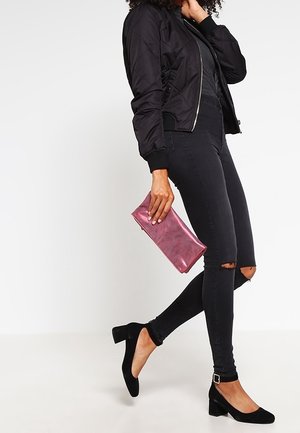 Giubbotto bomber nero trapuntato, jeans neri aderenti con dettagli strappati, scarpe con tacco a blocco in camoscio nero e una clutch metallica rosa.