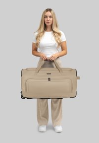Beige Reisetasche auf Rädern mit doppelten Tragegriffen, glatter Außenseite, Reißverschluss-Aufbewahrungstasche und schwarzen Hardware-Akzenten.