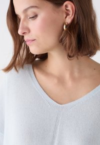 Maglione azzurro chiaro, morbido e a maglia, con scollo a V e dettagli cuciti discreti, abbinato a un semplice orecchino a cerchio dorato. Texture liscia.
