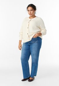 Kreemikas soonikkoes kardigan ümmarguse kaelusega ja nööpidega, kombineeritud siniste sirge säärega teksapükstega ja pruunide madalate kingadega.