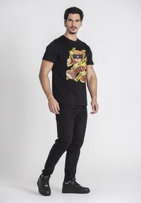 Mladý muž s tmavými vlasmi oblečený v čiernom tričku s potlačou medvedíka v slnečných okuliaroch obaleného žltou páskou, čiernych džínsach a teniskách.