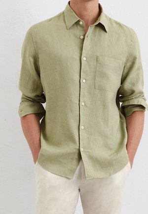 Mies, jolla on päällään vaaleanvihreä pitkähihainen nappipaita, hihat käärittyinä, ja beiget housut, kädet taskuissa, yksiväristä taustaa vasten.