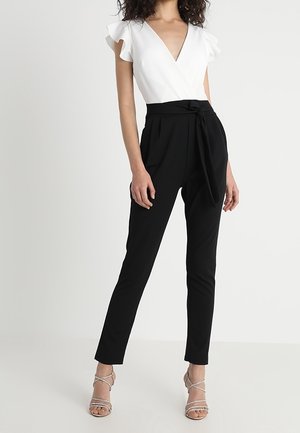 Nainen, joka pukeutunut valkoiseen liehuttavilla hihoilla varustettuun kietaisupaitaan, korkeavyötäröisiin mustiin housuihin, joissa on solmittava vyö, ja hopeisiin, remmillä varustettuihin korkokenkiin.