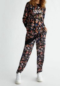 Květinová lounge souprava s černým základem, dlouhým rukávem s bílým logem a odpovídajícími kalhotami s živými květinovými vzory. Doplňena žebrovanými manžetami.