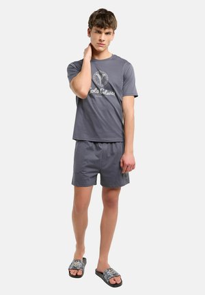 Jeune homme debout portant un t-shirt gris avec logo, un short assorti et des sandales noires à motifs et à bout ouvert, posant avec une main sur le cou.