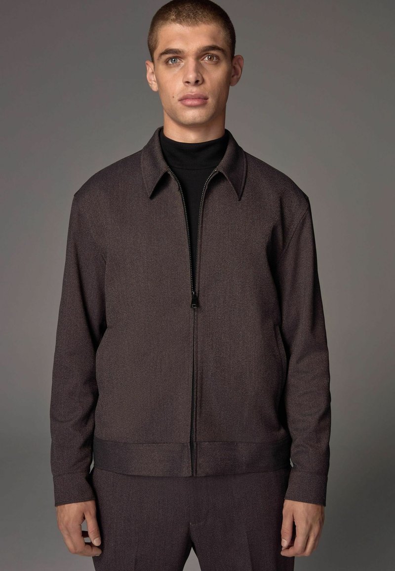 Dark brown zip-up jacket with a collared neckline, textured fabric, and side pockets. Paired with a matching pant.