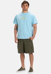 Lys blå bomull t-skjorte med grafisk logo, sammen med olivengrønne cargo-shorts med flere lommer. Grå sneakers fullfører antrekket.