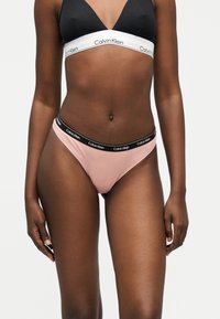 Γυναίκα που φοράει μαύρο τρίγωνο σουτιέν με λευκή ταινία Calvin Klein και ανοιχτό ροζ εσώρουχο στυλ μπικίνι με μαύρη ζώνη Calvin Klein.