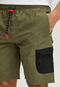 Oliven grønne shorts lavet af let materiale, med en sort netlomme med lynlås og justerbar snor i sort og rød.