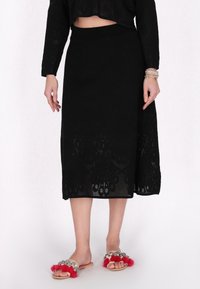 Fustă neagră de lungime medie, cu o textură asemănătoare crosetului și broderie florală la margine. Purtați cu sandale brodate, decorate cu ciucuri roșii.