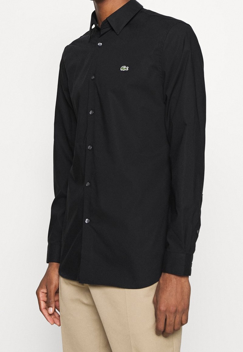 Camicia nera a maniche lunghe con bottoni, colletto a punta, logo verde sobrio sul petto, realizzata in tessuto morbido, dotata di bottoni neri.