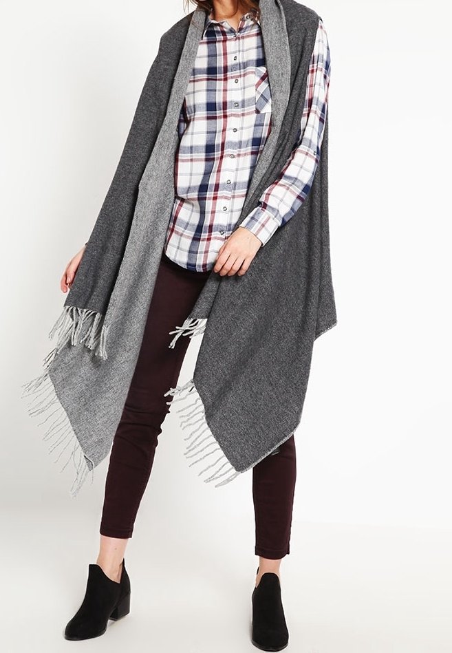Femme portant un châle gris à franges sur une chemise à carreaux, un pantalon foncé et des bottines noires, debout devant un fond blanc.