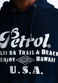 Marineblauwe hoodie met een voorzijde bedrukking in witte tekst. Kenmerken zijn onder andere verstelbare trekkoorden en een gestructureerde afwerking. De voorbedrukking bevat de merknaam en details.