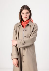 Beige Trenchcoat mit einem Knopffrontdesign, großem Kragen und hochgeschlagenen Manschetten. Kombiniert mit einem leuchtend roten Blumenschal. Glatte Textur.