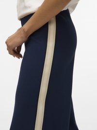 Vedere laterală a unei persoane purtând pantaloni bleumarin cu o dungă verticală lată bej și un top alb, cu mâna sprijinită aproape de șold.