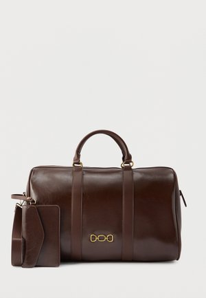 Σκούρο καφέ δερμάτινη τσάντα duffel με διπλές λαβές, έμβλημα αλυσίδας σε χρυσό και συνδεδεμένη δερμάτινη ετικέτα αποσκευών και λουράκι στο ίδιο χρώμα.