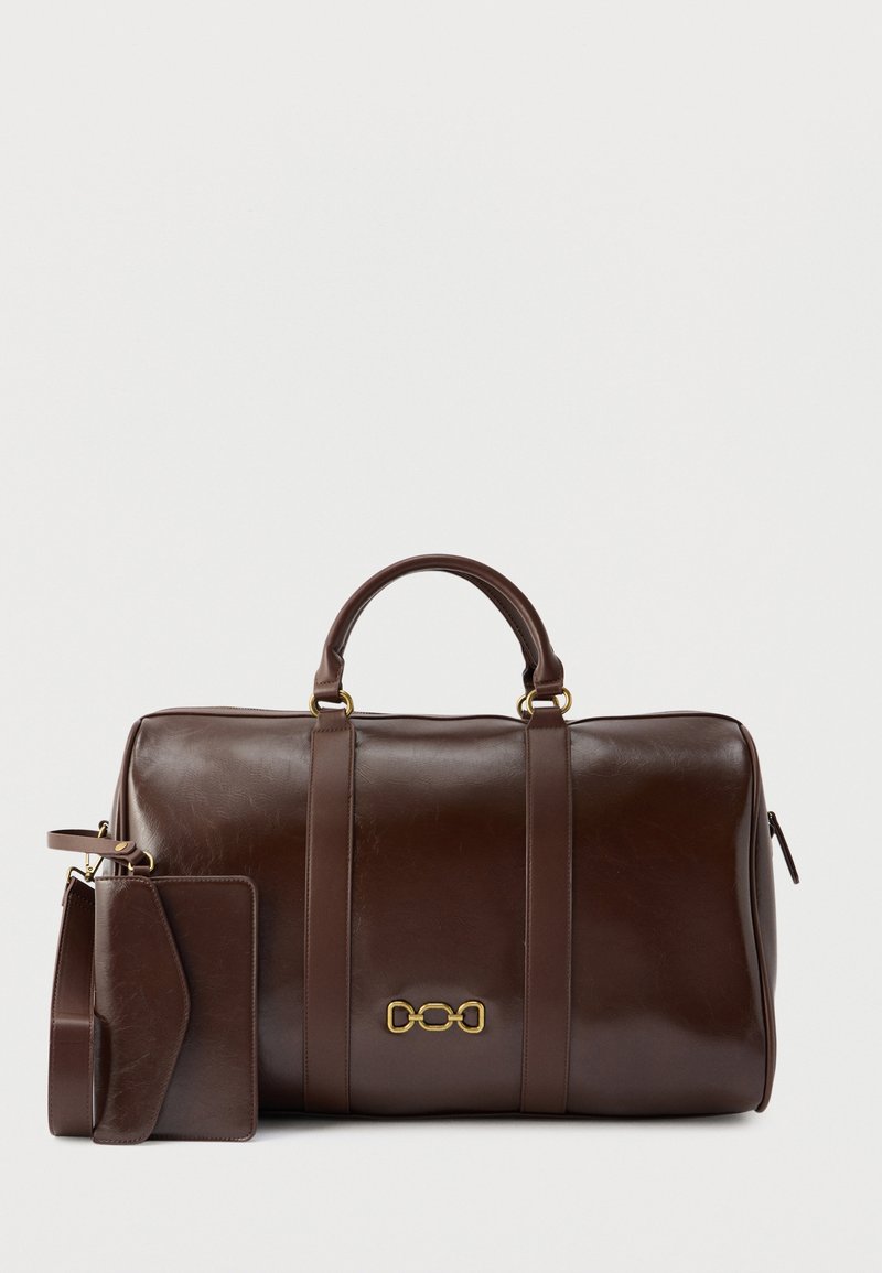 Σκούρο καφέ δερμάτινη τσάντα duffel με διπλές λαβές, έμβλημα αλυσίδας σε χρυσό και συνδεδεμένη δερμάτινη ετικέτα αποσκευών και λουράκι στο ίδιο χρώμα.