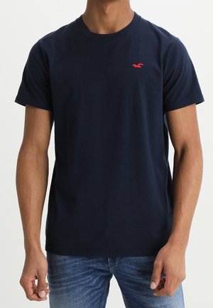 Mann trägt ein schlichtes marineblaues kurzärmeliges T-Shirt mit kleinem rotem Logo auf der linken Brust und blaue Jeans.