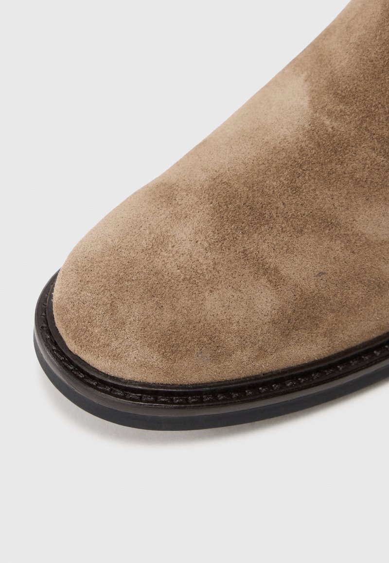 Beige Wildleder-Stiefelette mit einer runden Zehenform, ausgestattet mit einer schwarzen Gummisohle und dezenten Stichdetails entlang der Kante.