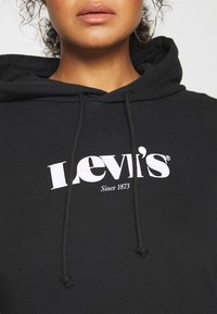 Primo piano di una persona che indossa una felpa nera Levi's con logo bianco e orecchini a cerchio dorati, labbra e mento visibili.