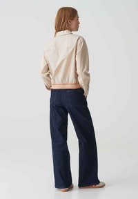 Lichtbeige jasje met elastische taille en contrasterende oranje details, gecombineerd met donkerblauwe wijde jeans, gezien vanuit de achterkant.