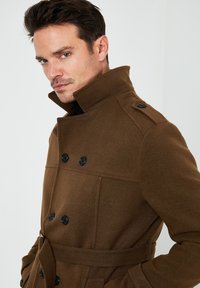 Abrigo de lana marrón con cuello alto, cierre de botones doble y cinturón. Presenta botones negros y un acabado suave y texturizado.