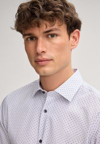 Chemise blanche avec un petit motif géométrique, dotée d'un col pointu et de boutons foncés, fabriquée dans un tissu lisse avec une texture légère.