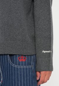 Close-up van een hand dichtbij de grijze gebreide mouw van een trui met de tekst "Originals" en een spijkerbroek met blauwe streepjes en een rode geborduurde kroon op de zak.