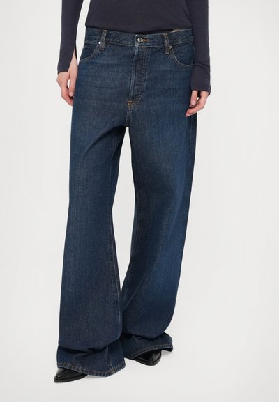 Jean en denim bleu foncé à jambes larges, taille haute, poches avant et détails de surpiqûres visibles. Le tissu a une texture légère et lisse.