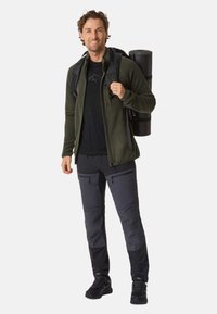 Giacca outdoor verde oliva, t-shirt nera, pantaloni grigio scuro con tasche con zip, scarpe nere, che trasporta un tappetino da yoga nero nello zaino.