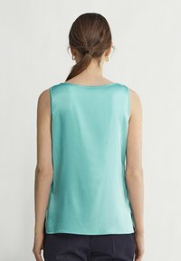 En ärmlös, ljus turkos satinblus med rundad halslinning och sidofickor, med en jämn textur och en minimalistisk design.