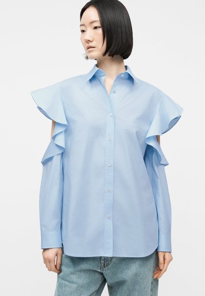 Femme aux cheveux noirs courts portant un chemisier bleu clair à boutons avec des manches épaules dénudées à volants et un jean bleu clair.