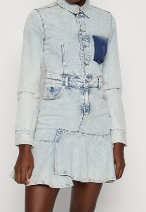 Femme portant une robe en jean bleu clair à manches longues, boutonnée sur le devant, poche poitrine bleu foncé et détail de jupe évasée.