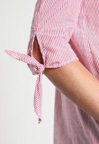 Maglietta a maniche corte a righe rosse e bianche con un dettaglio a fiocco annodato sulla manica, posizionata sopra l'avambraccio di una persona su uno sfondo neutro.