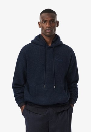 Man in een donkerblauwe hoodie over een zwart shirt, staand met zijn handen in de zakken tegen een effen witte achtergrond.
