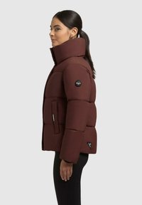 Donna di profilo che indossa un giubbotto imbottito marrone scuro con colletto alto, tasche laterali e leggings neri su uno sfondo chiaro e semplice.