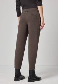 Bruine taps toelopende broek met een gladde textuur, elastische tailleband en twee achterzakken. Gecombineerd met zwarte enkellaarsjes.