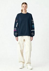 Jūras spēku pelēks džemperis ar dažādiem krāsainiem izšūtiem motīviem uz piedurknēm, saskaņots ar brīviem baltiem cargo biksēm un masīviem baltajiem sporta apaviem.