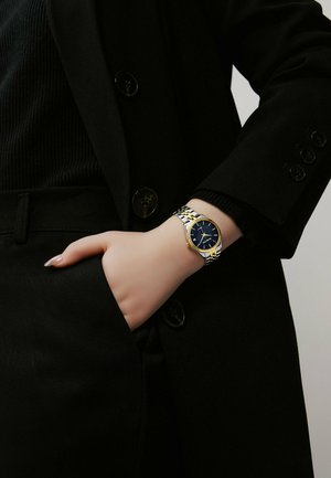Orologio d'oro e argento con quadrante blu, bracciale in acciaio inossidabile e dettagli dorati, indossato su un polso contro un outfit nero.