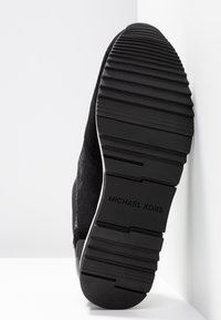 Semelle noire d'une chaussure Michael Kors montrant une bande de roulement texturée et le nom de la marque embossé au centre sur un fond blanc.