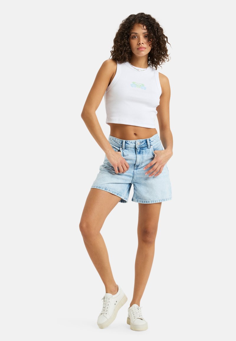 Witte cropped tanktop met een lichtgroene grafische print, gecombineerd met lichtblauwe denim shorts. Model draagt witte sneakers. Eenvoudig en casual ontwerp.