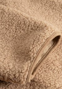 Bež flis tkanina s teksturiranom, pahuljastom površinom i glatkim obrubom. Ima mekan, topao osjećaj s vidljivim šavovima i prianjajućim rubom.