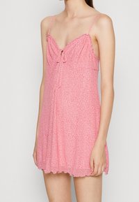 Růžové květinové šaty s tenkými ramínky, řasenými okraji a mašlí na výstřihu. Lehká látka s jemnou strukturou a rozšířeným střihem.