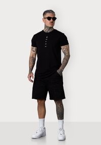 Schwarzes T-Shirt mit vertikalem Text, schwarze Cargo-Shorts, weiße Turnschuhe und Sonnenbrille. Tätowierungen sichtbar an Armen und Beinen. Minimalistisches Design.