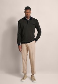 Dunkelgrüner Pullover mit einem marineblauen Kragen, kombiniert mit hellbeigen Hosen und beigen Sneakers. Auffällige Brusttasche und gerippte Bündchen.