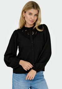 Giovane donna con capelli biondi indossa una blouse nera a maniche lunghe con collare in pizzo e maniche a sbuffo, abbinata a jeans blu chiaro.