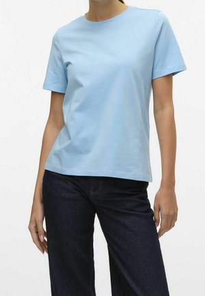Kobieta ubrana w jasnoniebieski T-shirt z krótkim rękawem i okrągłym dekoltem oraz ciemnoniebieskie jeansy, stojąca na tle jednolitego jasnego tła.